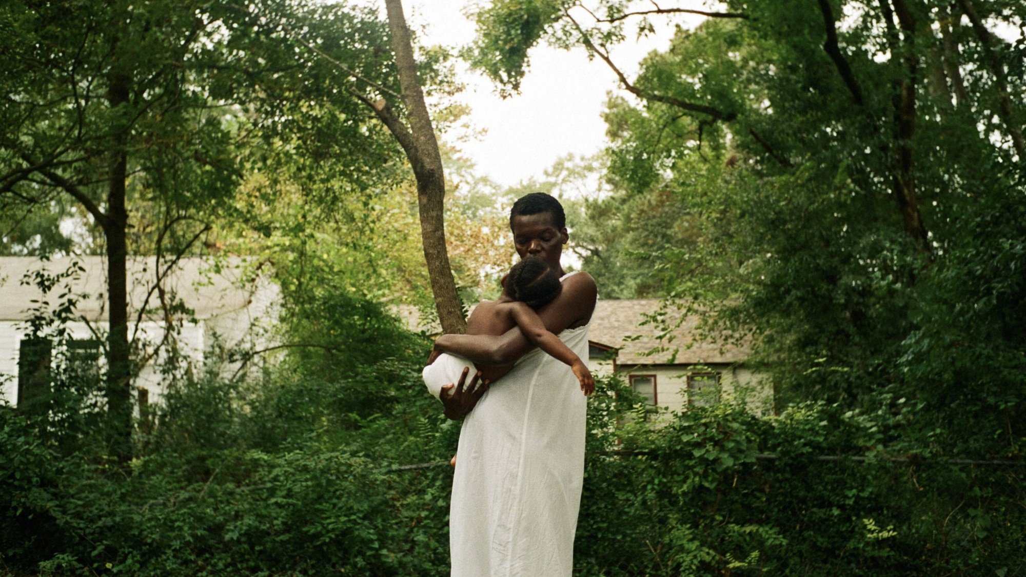A woman in a white dress holds her baby while standing outside among green trees.